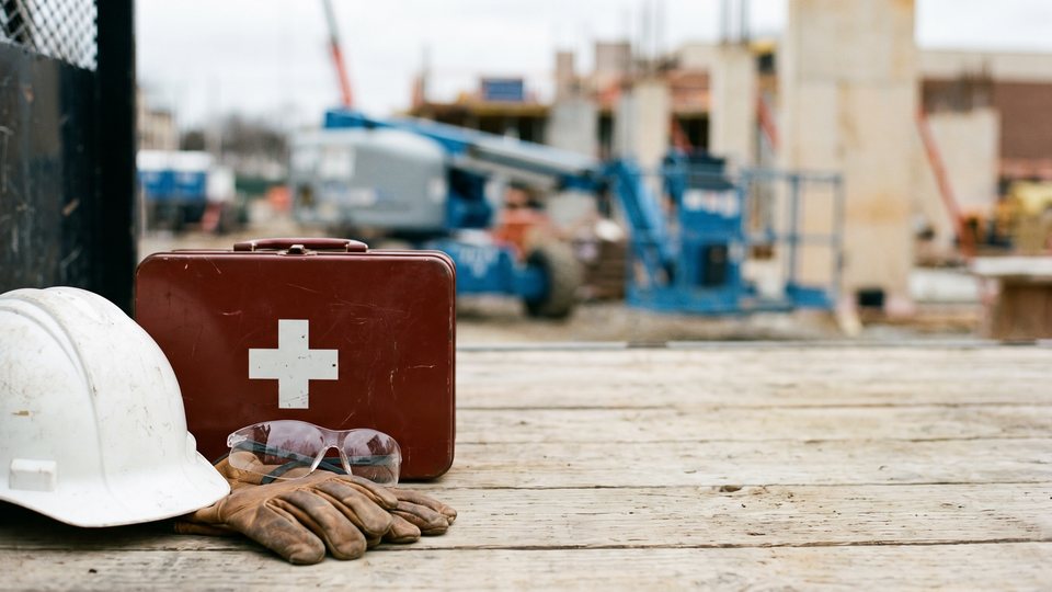 A white hard hat, work gloves, safety glasses and a red first-aid case on a tailgate at a busy jobsite