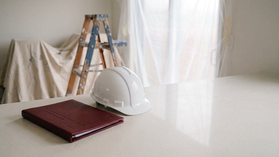 A red document folder and a white hard hat on a kitchen countertop during a remodel, with drop cloths and a step ladder behind